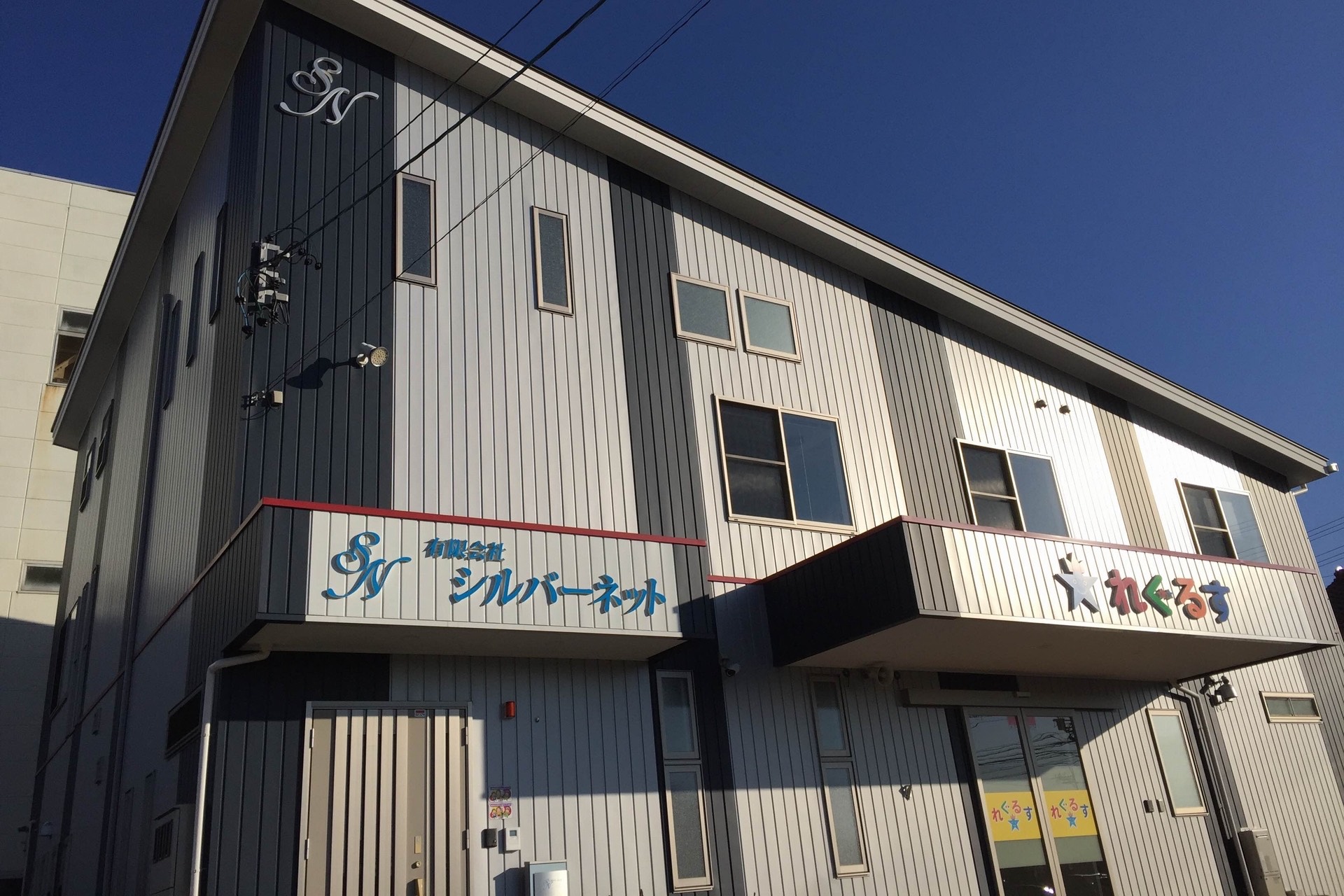 介護施設の建物外観。白と黒の縦張りサイディングの2階建て、複数の窓と施設看板が見える。